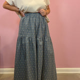 A person wearing a cream short-sleeve top and a high-waisted, tiered blue-gray wide-leg skirt with a small white geometric pattern, standing against a pink wall.