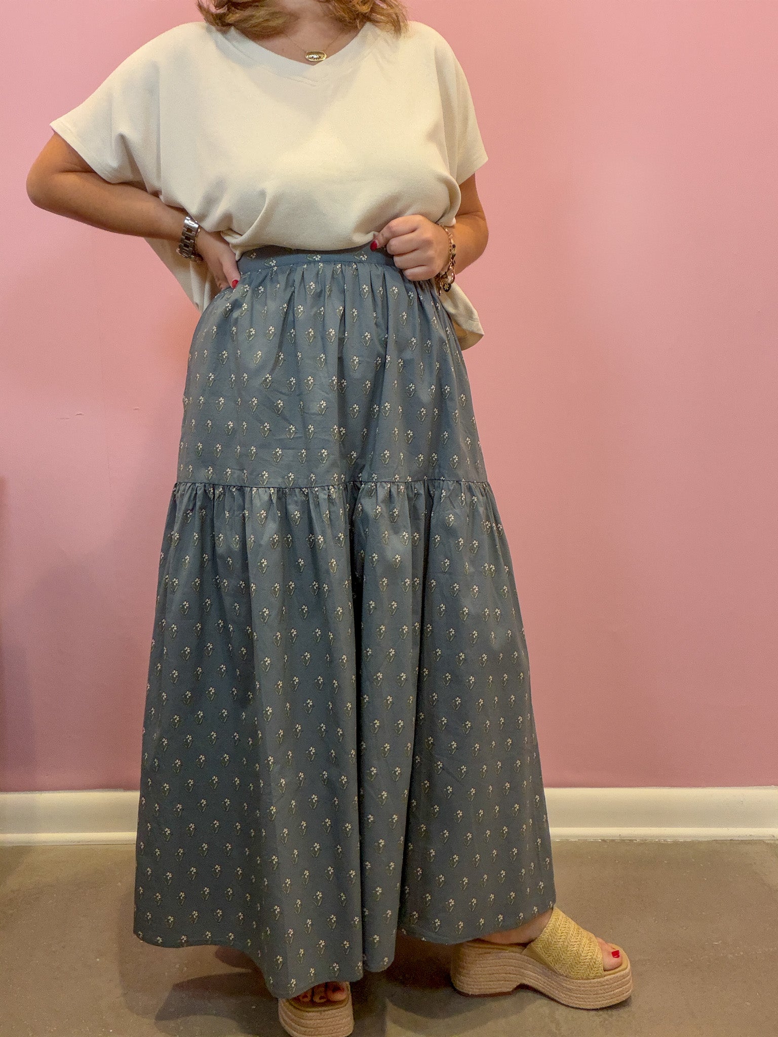 A person wearing a cream short-sleeve top and a high-waisted, tiered blue-gray wide-leg skirt with a small white geometric pattern, standing against a pink wall.