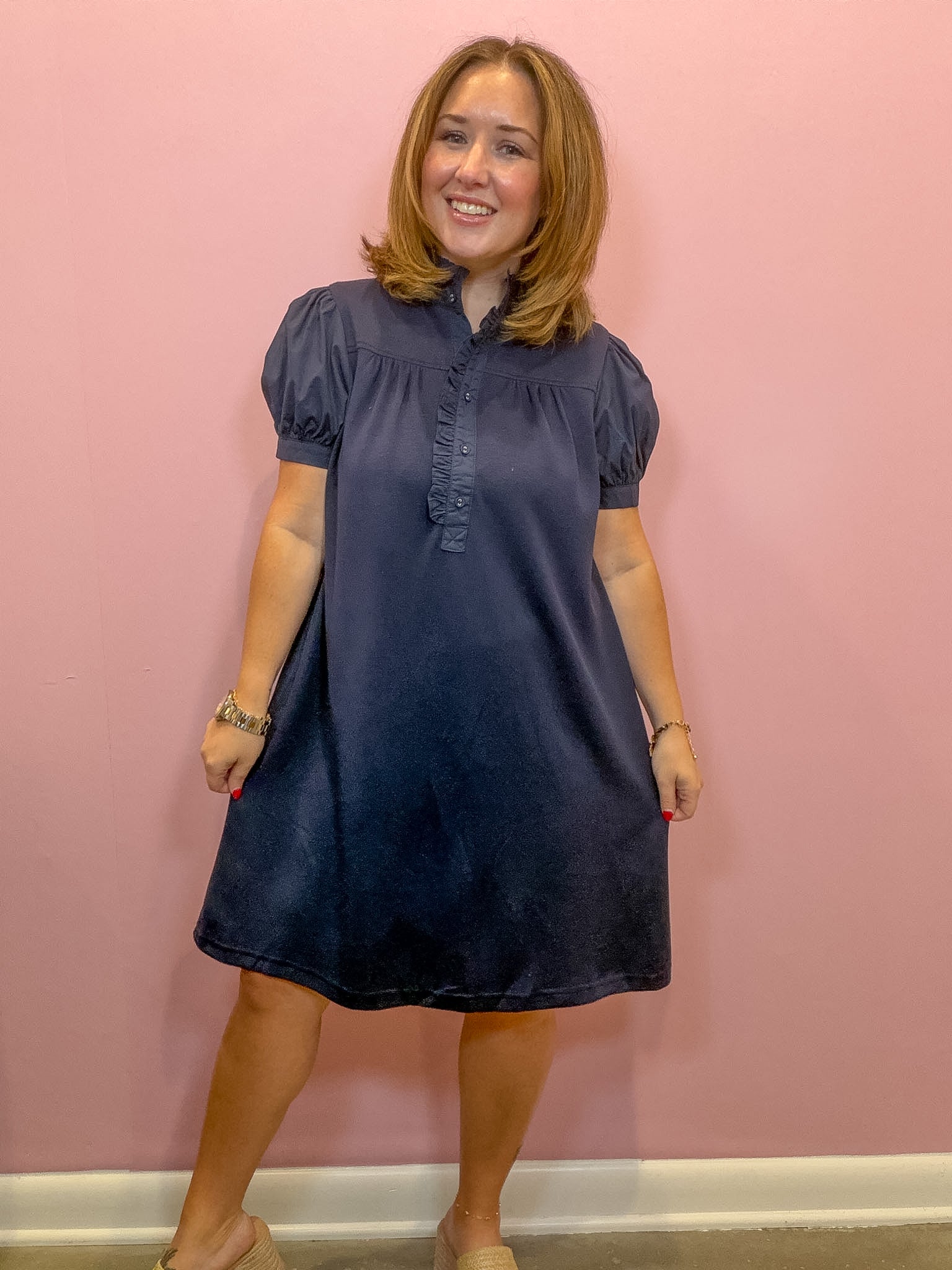A woman modeling a dark navy blue short-sleeve dress with puff sleeves and a ruffled collar, standing against a solid pink wall.