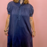 A woman wearing a navy blue short-sleeve shift dress with a ruffled button placket, standing against a solid pink wall.
