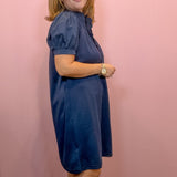 A woman modeling a navy blue short-sleeve dress with puff sleeves and a ruffled neckline, standing against a solid pink wall.