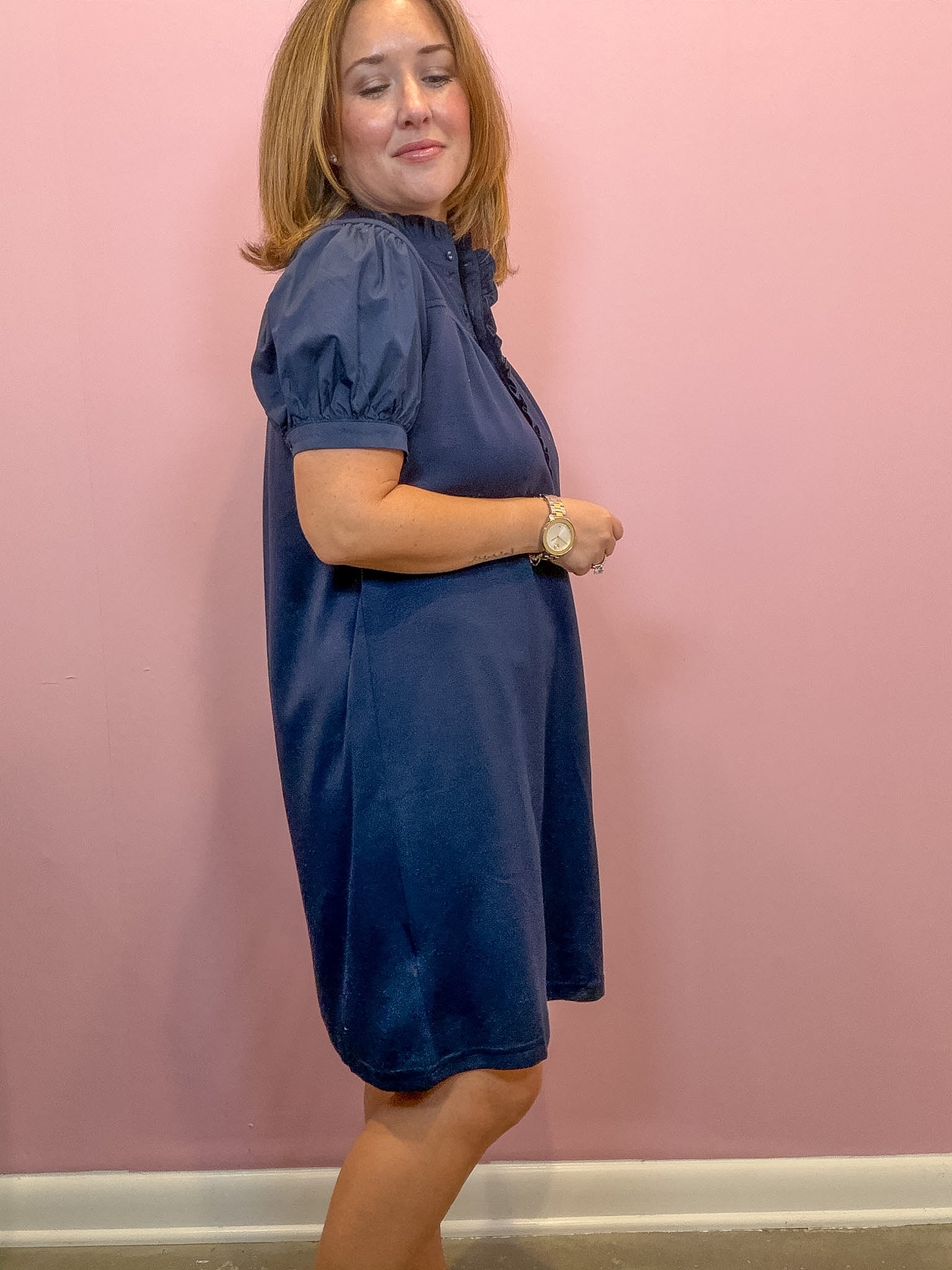 A woman modeling a navy blue short-sleeve dress with puff sleeves and a ruffled neckline, standing against a solid pink wall.