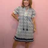 A woman modeling a short-sleeve tunic dress with a navy and white geometric pattern, standing against a solid pink wall.