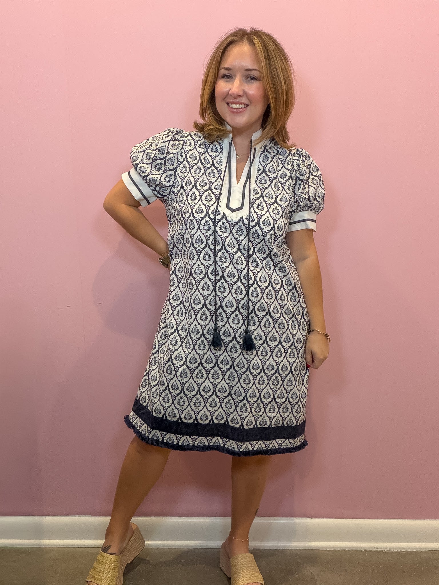 A woman modeling a short-sleeve tunic dress with a navy and white geometric pattern, standing against a solid pink wall.