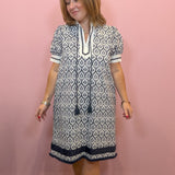 A woman modeling a short-sleeve tunic dress with a navy and white geometric pattern, standing against a solid pink wall.