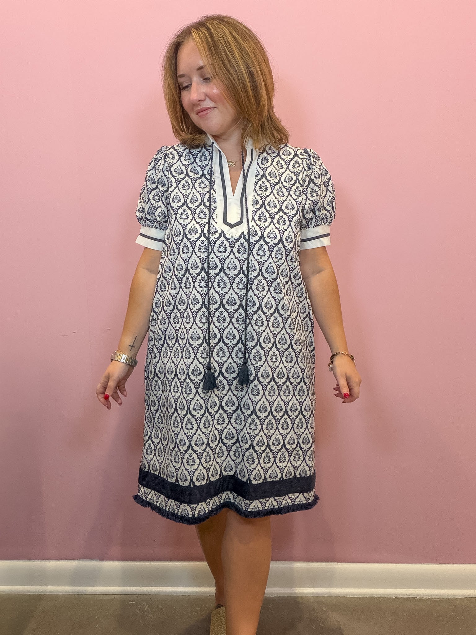 A woman modeling a short-sleeve tunic dress with a navy and white geometric pattern, standing against a solid pink wall.