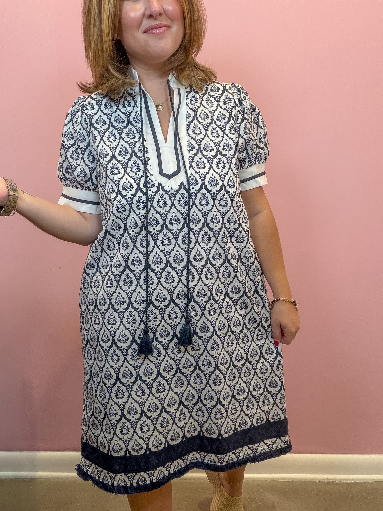 A woman modeling a short-sleeve tunic dress with a navy and white geometric pattern, standing against a solid pink wall.