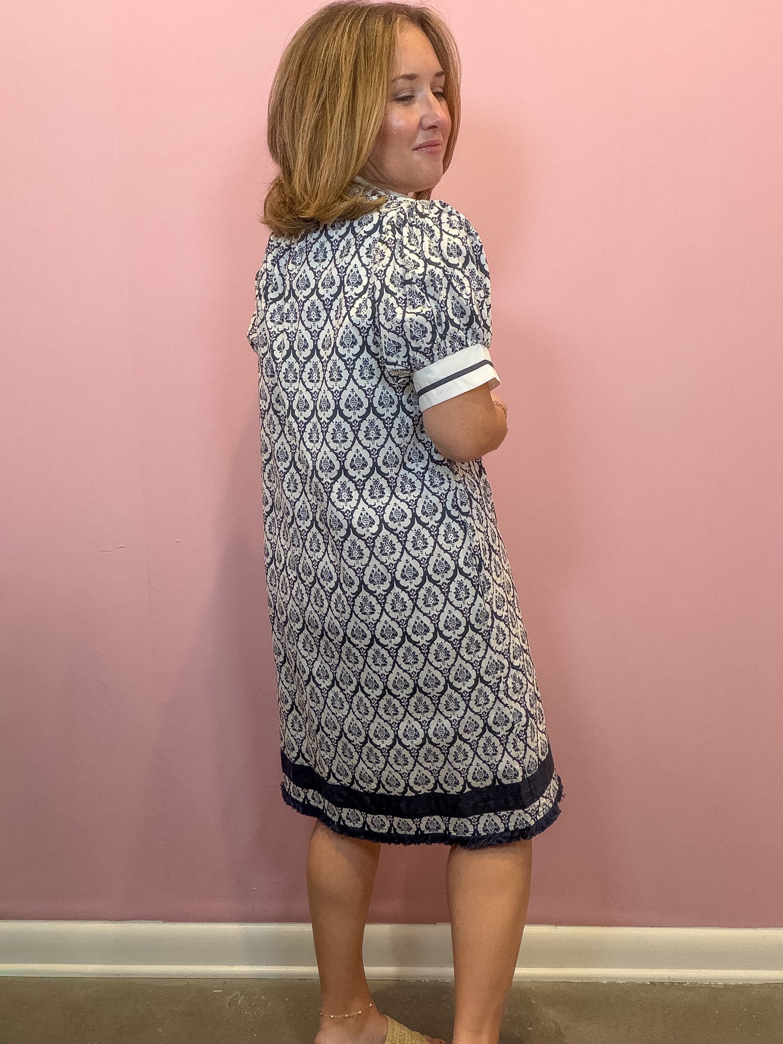 A woman wearing a short-sleeve navy and white patterned dress, standing against a solid pink wall.