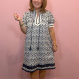 A woman modeling a short-sleeve tunic dress with a navy blue and white geometric block print pattern, standing against a solid pink wall.