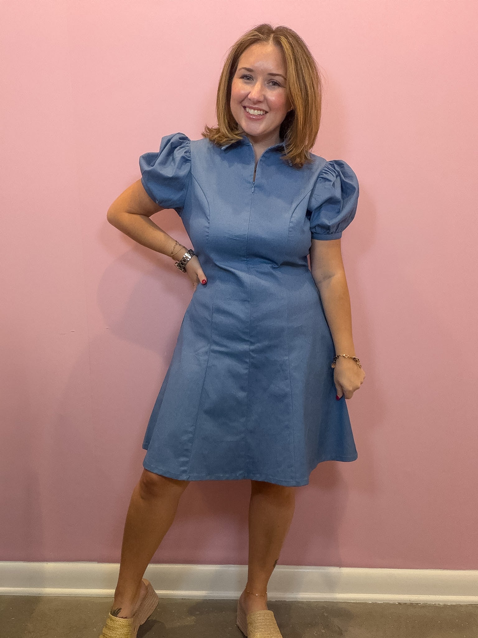 A woman modeling a blue short-sleeve dress with puff sleeves and a high mock neck, standing against a solid pink wall.