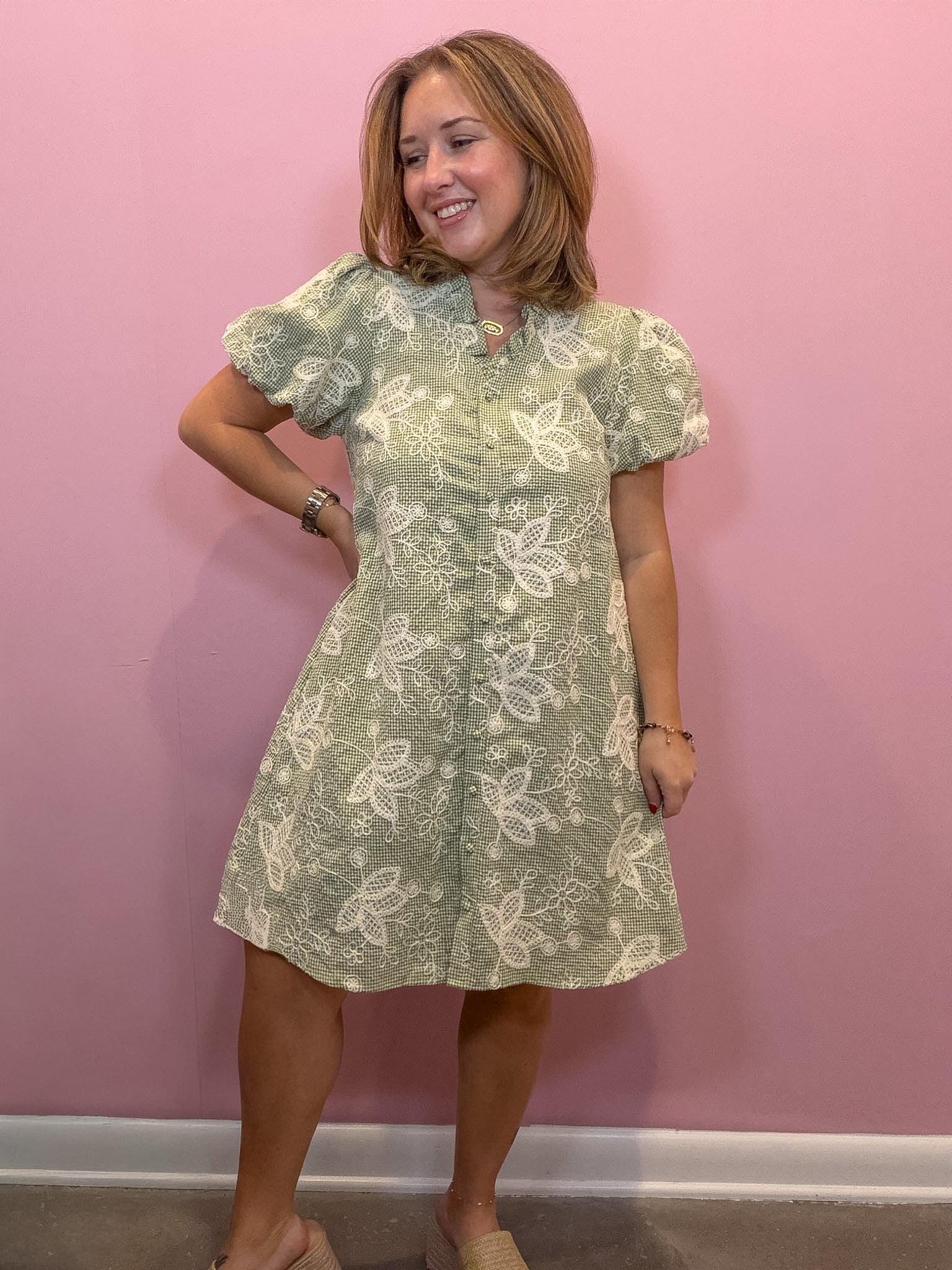 A woman modeling a light sage green and white butterfly-patterned short-sleeve dress against a solid pink wall.