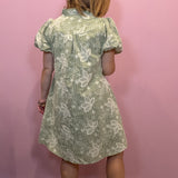 A child wearing a short-sleeve dress with a white dragonfly pattern on a sage green background, shown from the back against a pink wall.