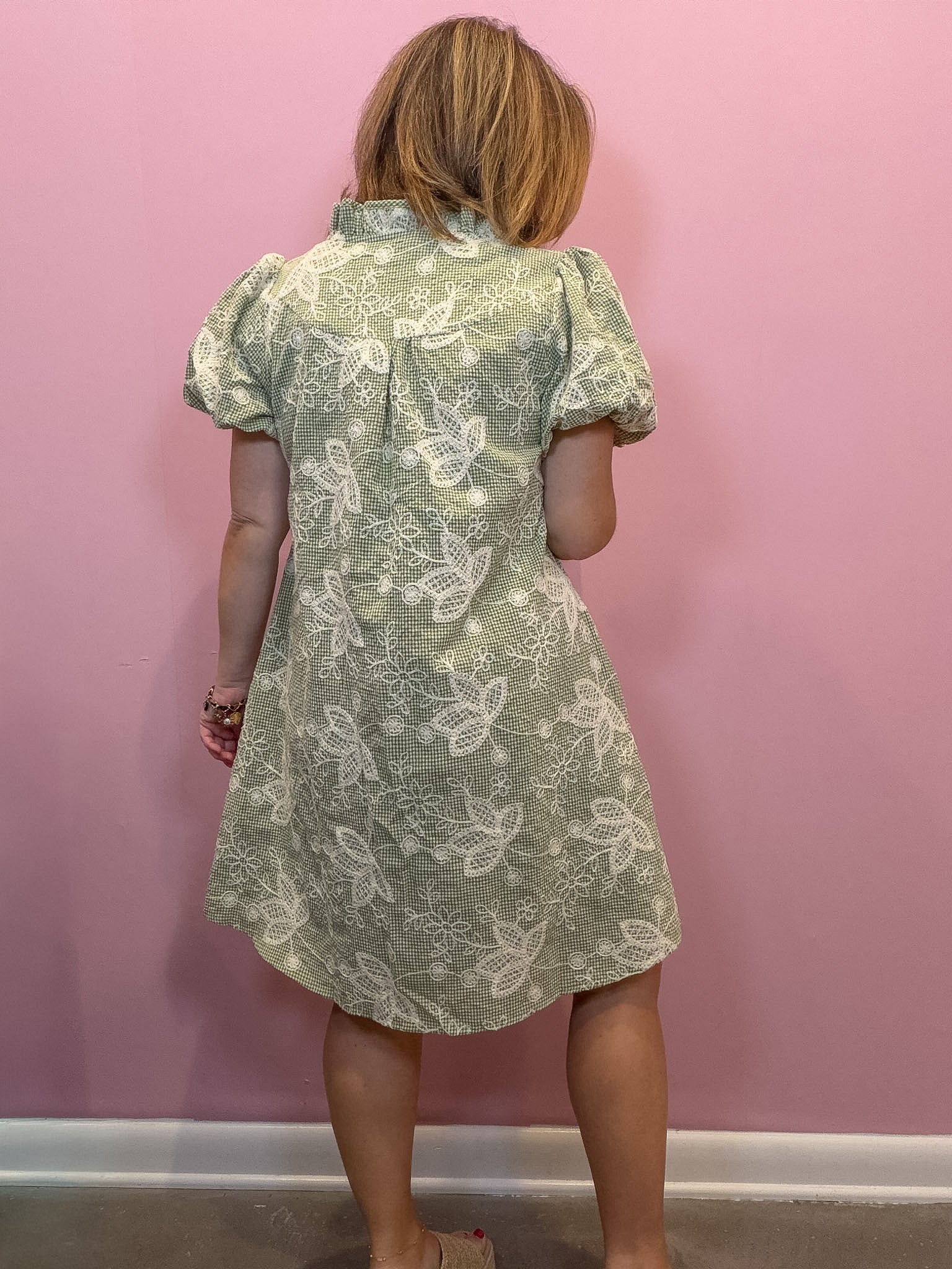A child wearing a short-sleeve dress with a white dragonfly pattern on a sage green background, shown from the back against a pink wall.