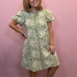 A woman modeling a sage green and white floral print puff-sleeve shift dress, standing against a solid pink wall.