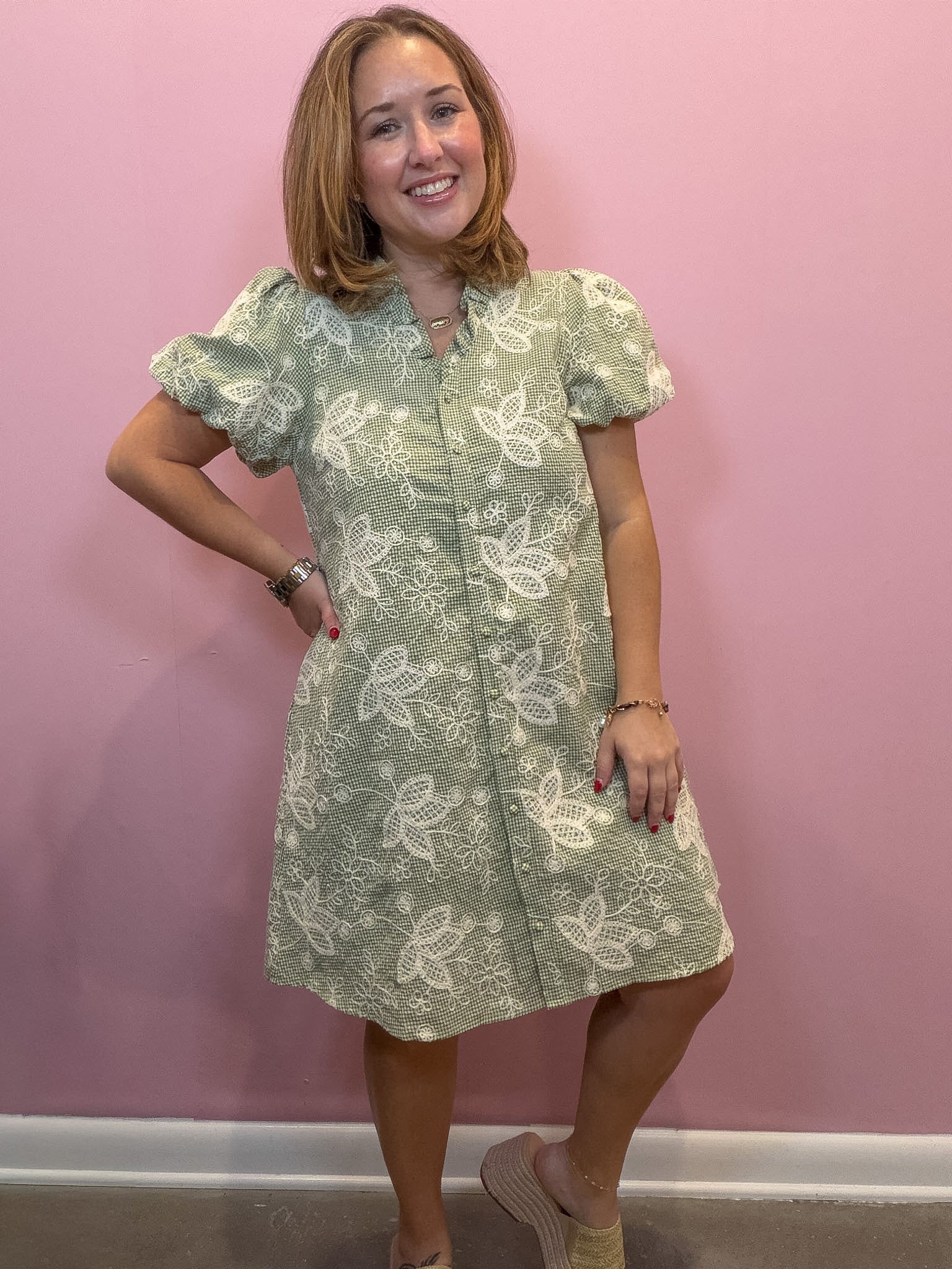 A woman modeling a sage green and white floral print puff-sleeve shift dress, standing against a solid pink wall.