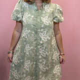 A woman wearing a sage green and white floral lace dress with puff sleeves and a button-up front, standing against a solid pink wall.