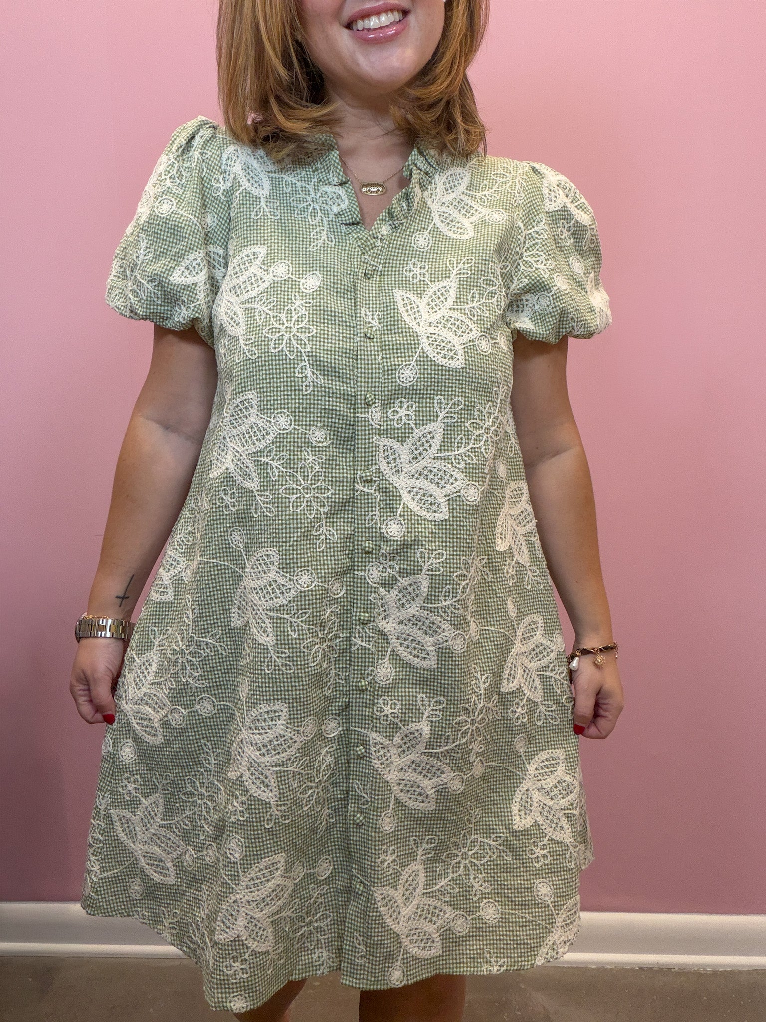 A woman wearing a sage green and white floral lace dress with puff sleeves and a button-up front, standing against a solid pink wall.