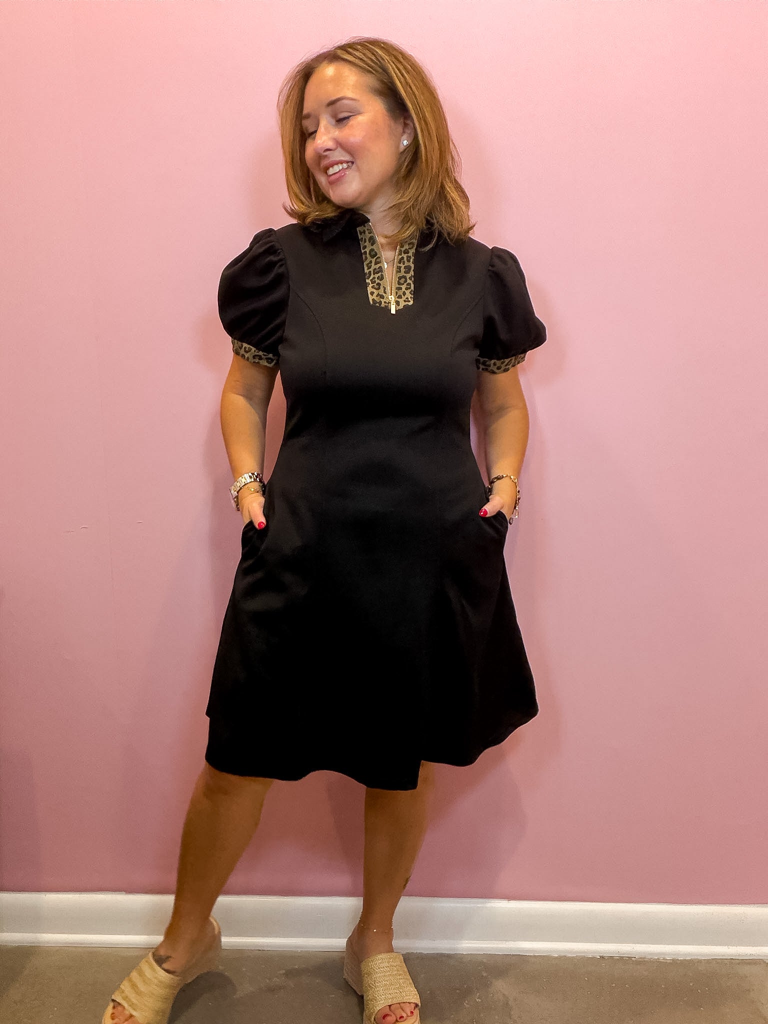 A woman modeling a black short-sleeve dress with gold patterned trim at the collar and sleeve cuffs, standing against a solid pink wall.