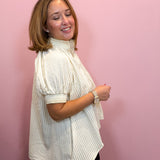 A woman with shoulder-length brown hair models a cream-colored, vertically striped short-sleeve blouse with puff sleeves, paired with black bottoms, standing against a solid pink wall.