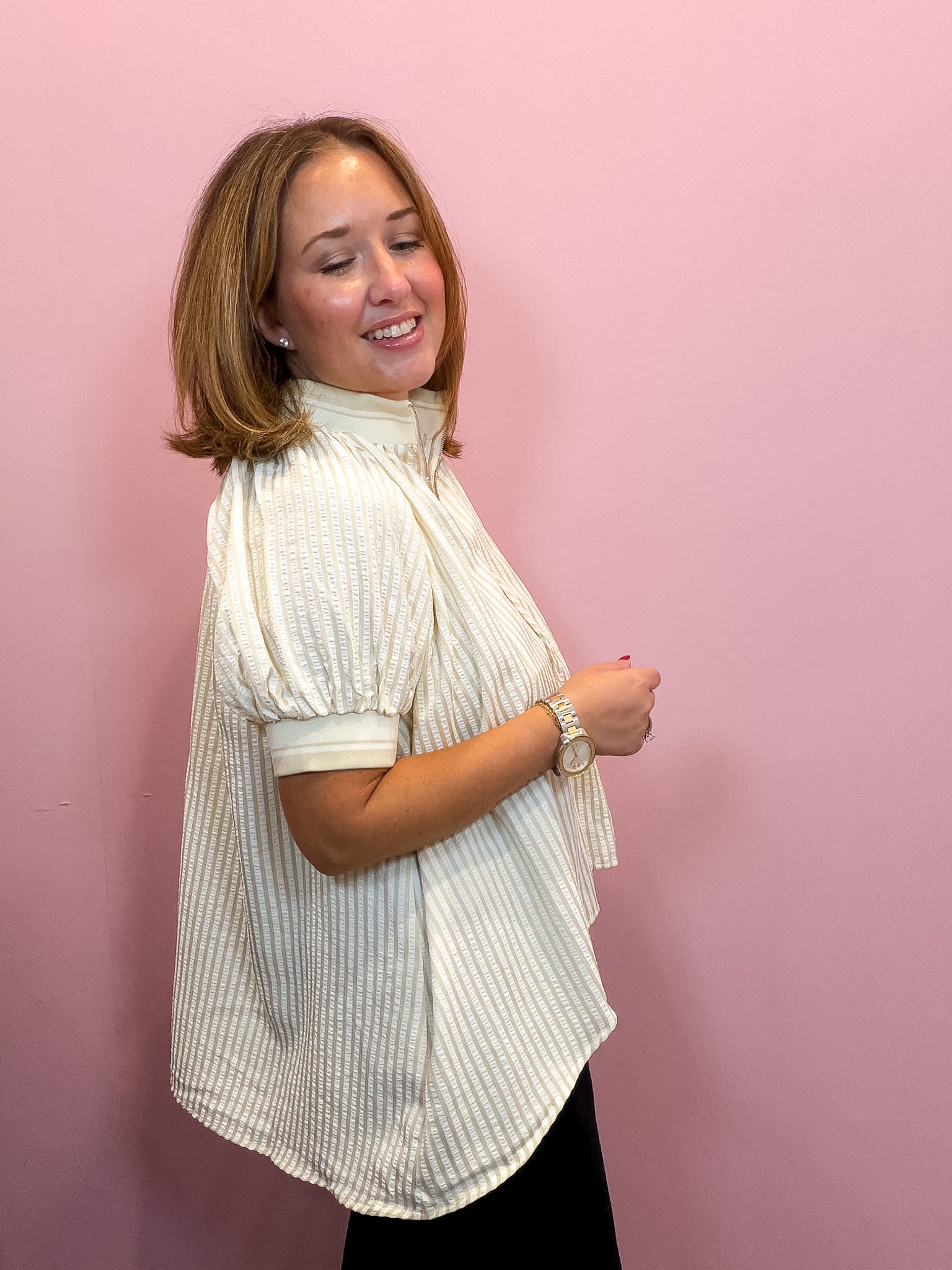 A woman with shoulder-length brown hair models a cream-colored, vertically striped short-sleeve blouse with puff sleeves, paired with black bottoms, standing against a solid pink wall.