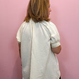 A woman viewed from the back wearing a cream and white vertically striped short-sleeve blouse with puff sleeves, set against a solid pink wall.