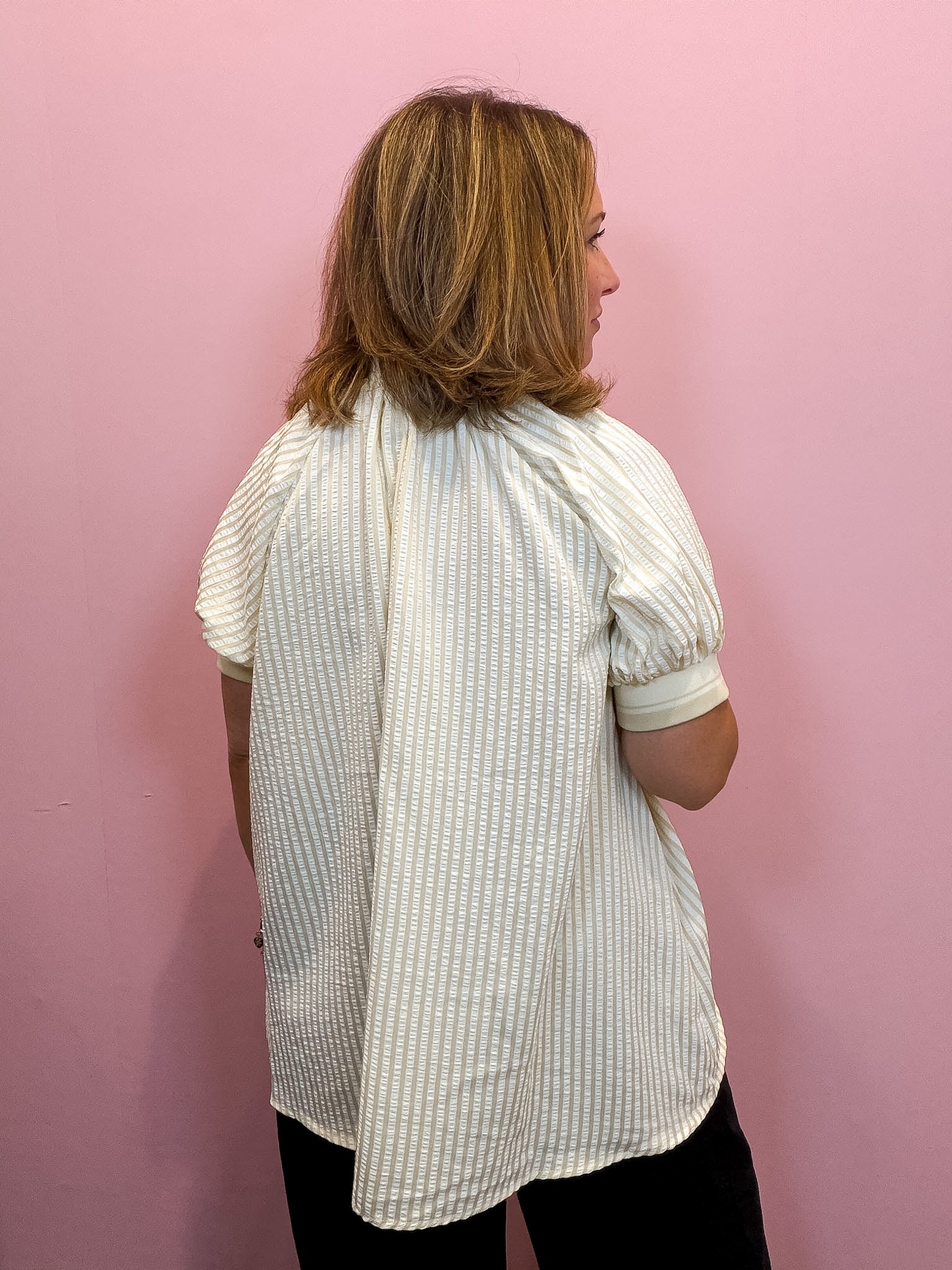 A woman viewed from the back wearing a cream and white vertically striped short-sleeve blouse with puff sleeves, set against a solid pink wall.