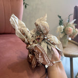 A hand holding a small gift box wrapped in a beige and brown patterned silk scarf, set against a soft pink velvet armchair in a cozy indoor setting.