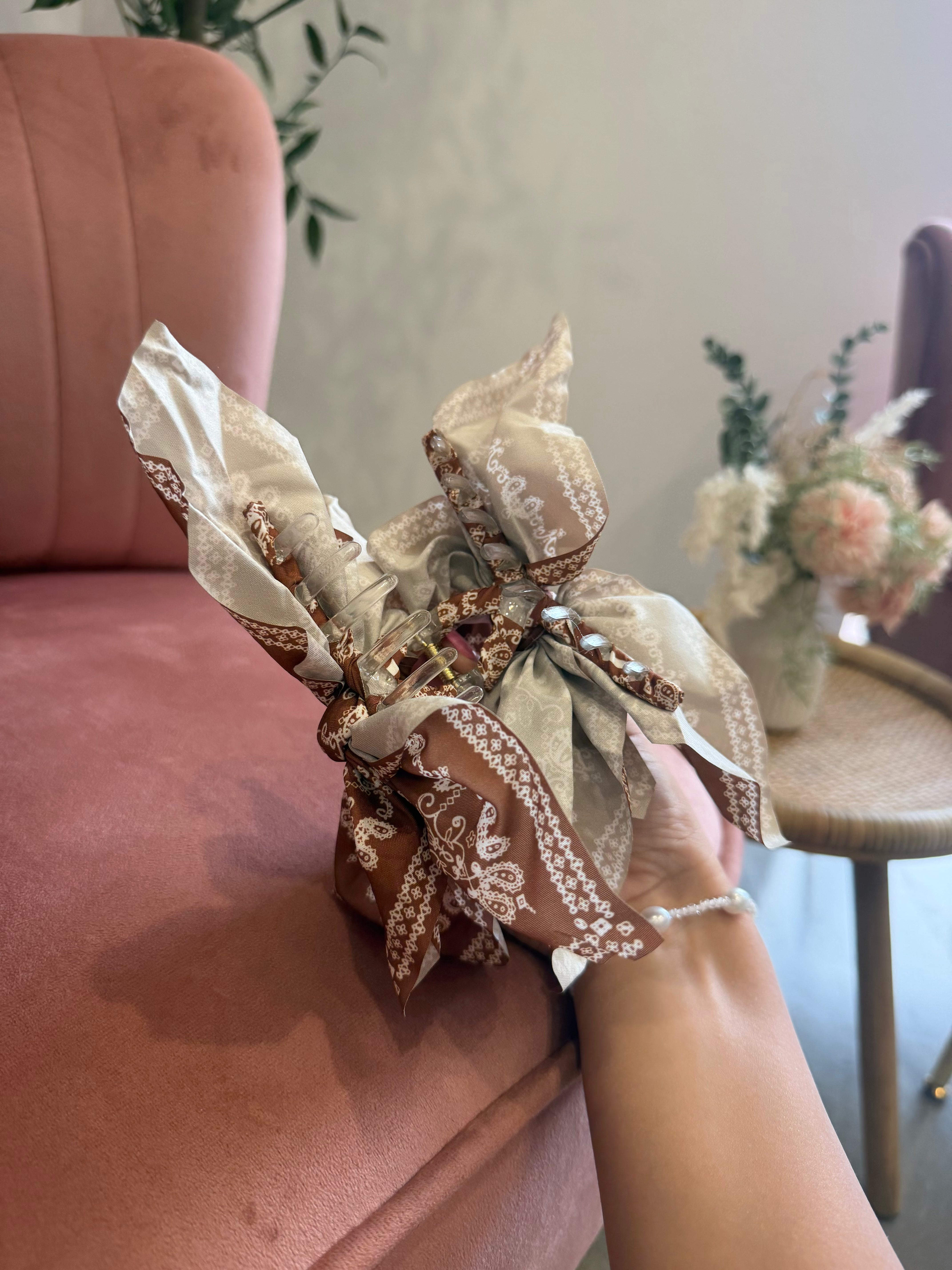 A hand holding a small gift box wrapped in a beige and brown patterned silk scarf, set against a soft pink velvet armchair in a cozy indoor setting.
