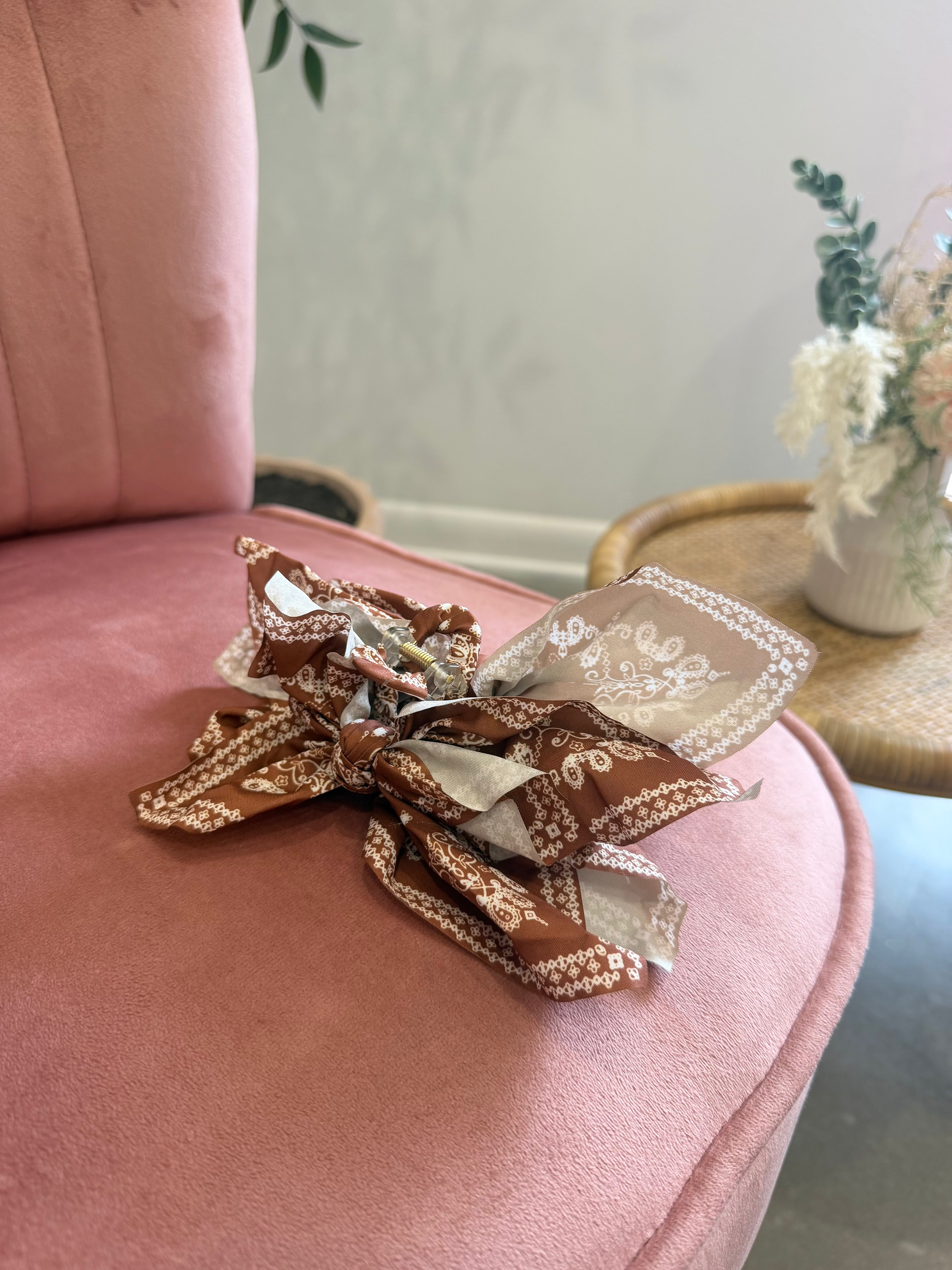 A decorative hair bow made from patterned fabric bands in brown, beige, and white, resting on a dusty pink velvet armchair in a softly styled indoor setting.