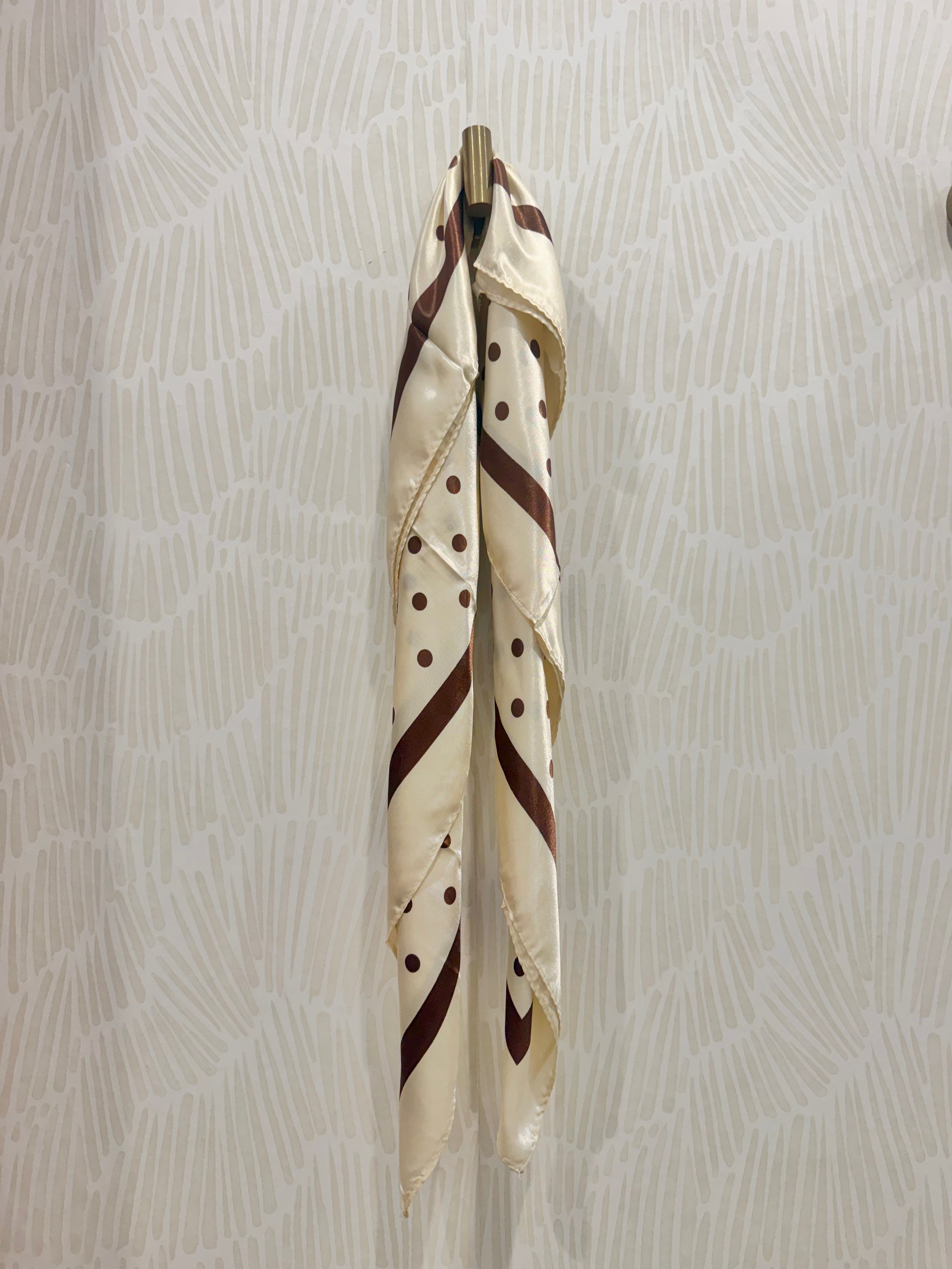 A cream-colored silk scarf with brown stripes, polka dots, and a diagonal stripe pattern, hanging from a gold metal ring against a light beige patterned wallpaper background.