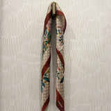 A patterned silk scarf with a beige and white checkered base, red stripes, and green floral motifs, hanging from a brass hook on a textured off-white wall.