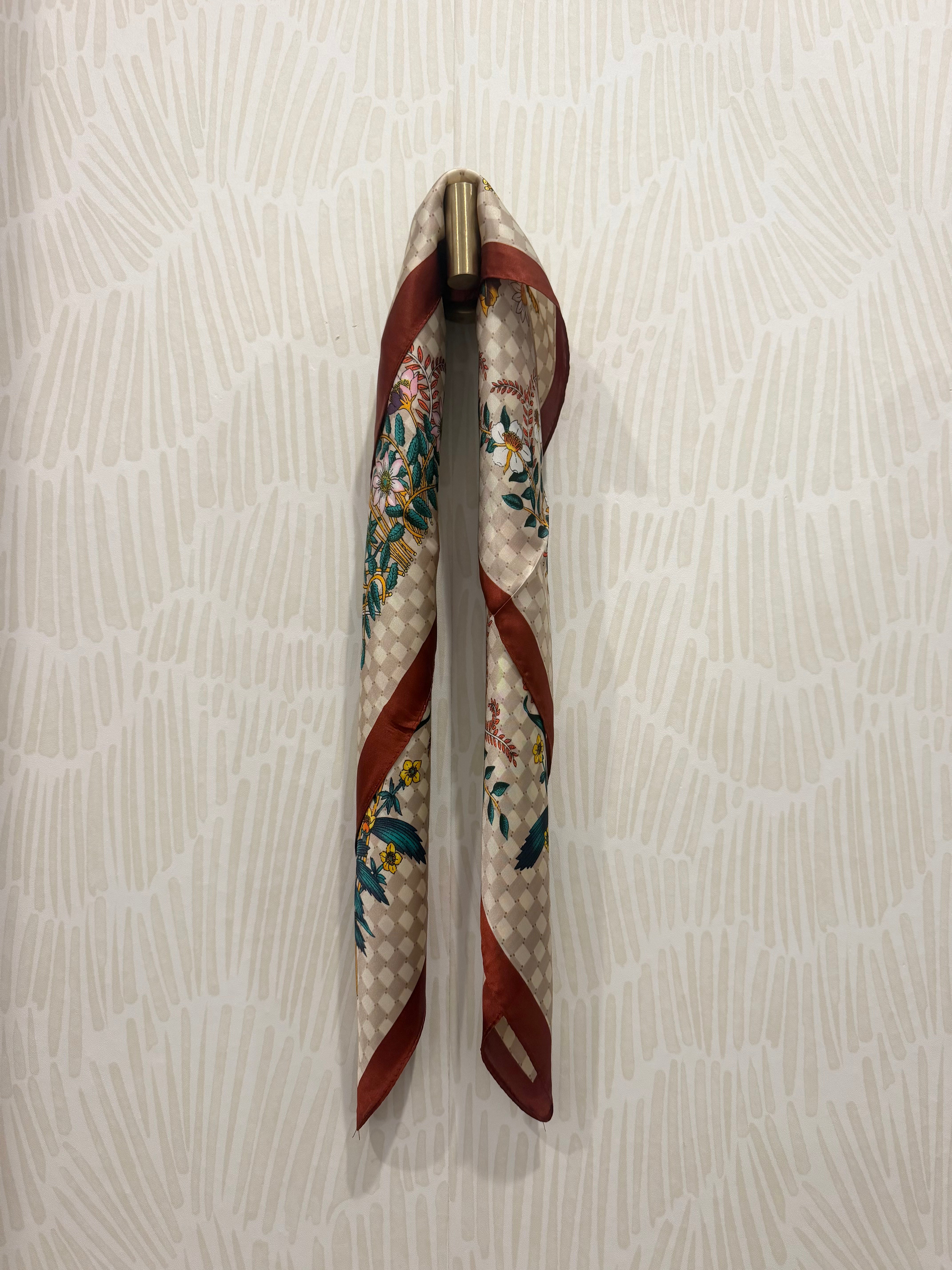 A patterned silk scarf with a beige and white checkered base, red stripes, and green floral motifs, hanging from a brass hook on a textured off-white wall.