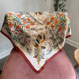 A square scarf with a cream base, orange floral print, and green and blue botanical details, draped over a dusty pink velvet armchair in an indoor setting.