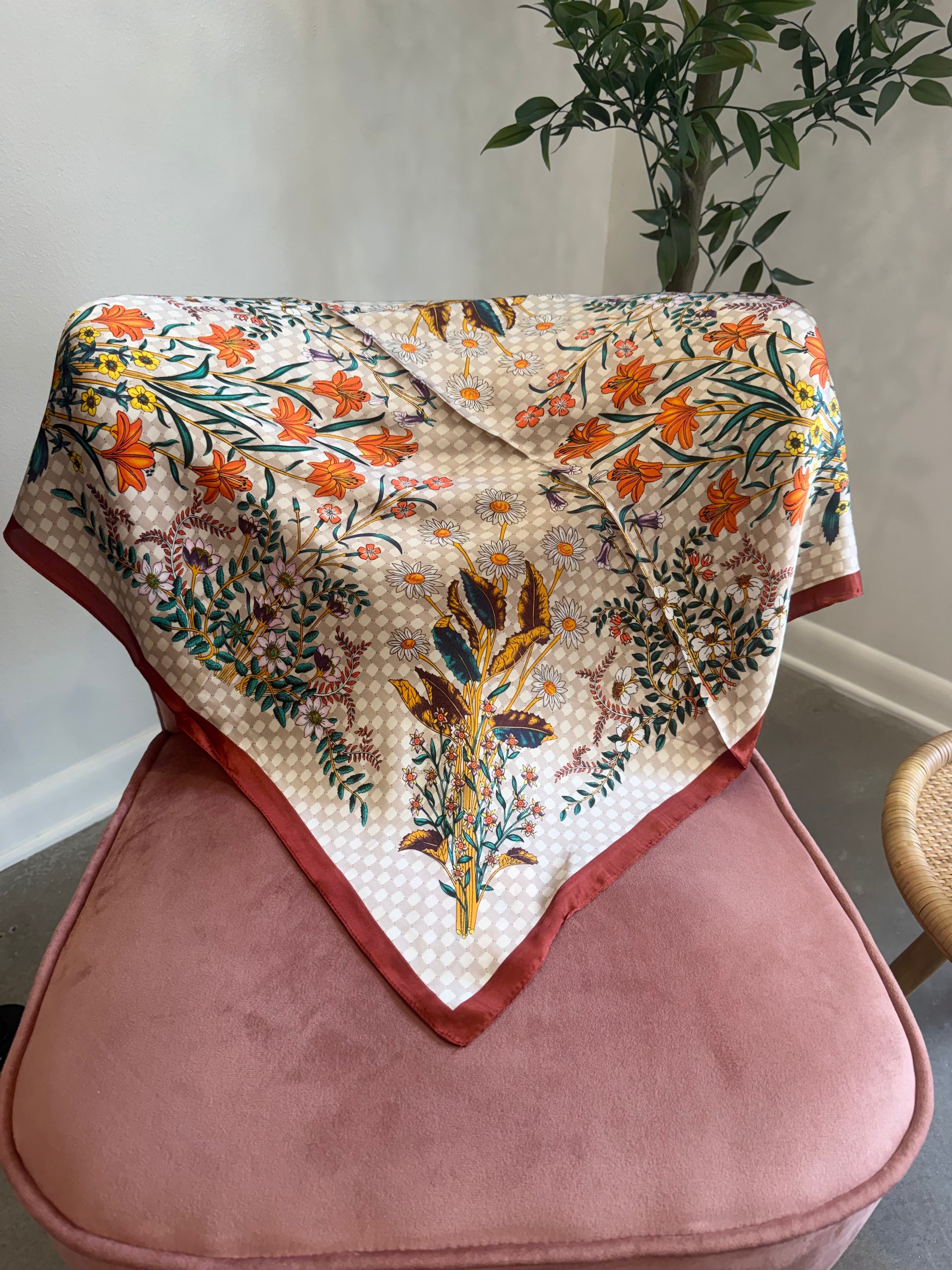 A square scarf with a cream base, orange floral print, and green and blue botanical details, draped over a dusty pink velvet armchair in an indoor setting.