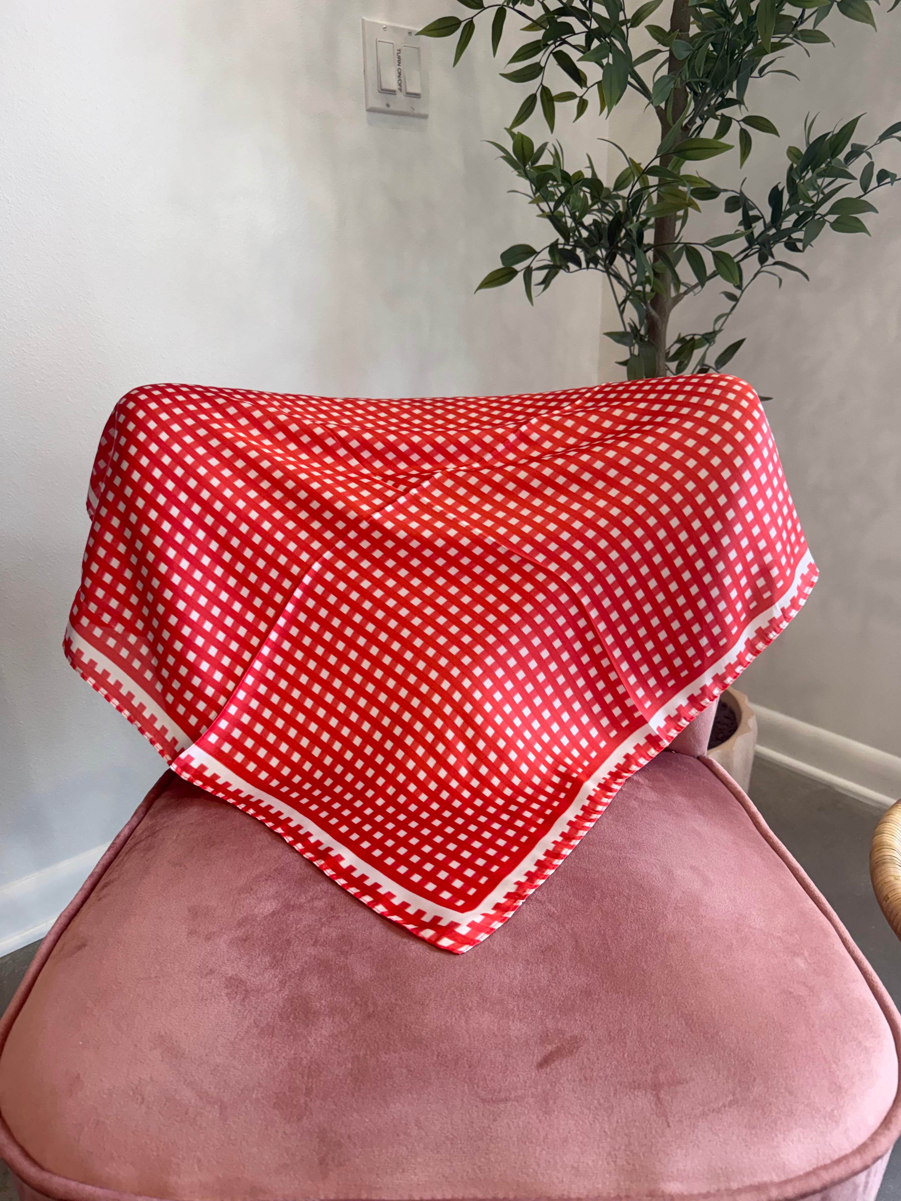 A red and white gingham checkered bandana or scarf draped over a dusty pink velvet armchair in a home setting, with a potted plant and wall visible in the background.