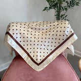 A cream-colored square scarf with brown polka dots and a brown border, draped over a dusty pink velvet chair in an indoor setting with a potted plant in the background.