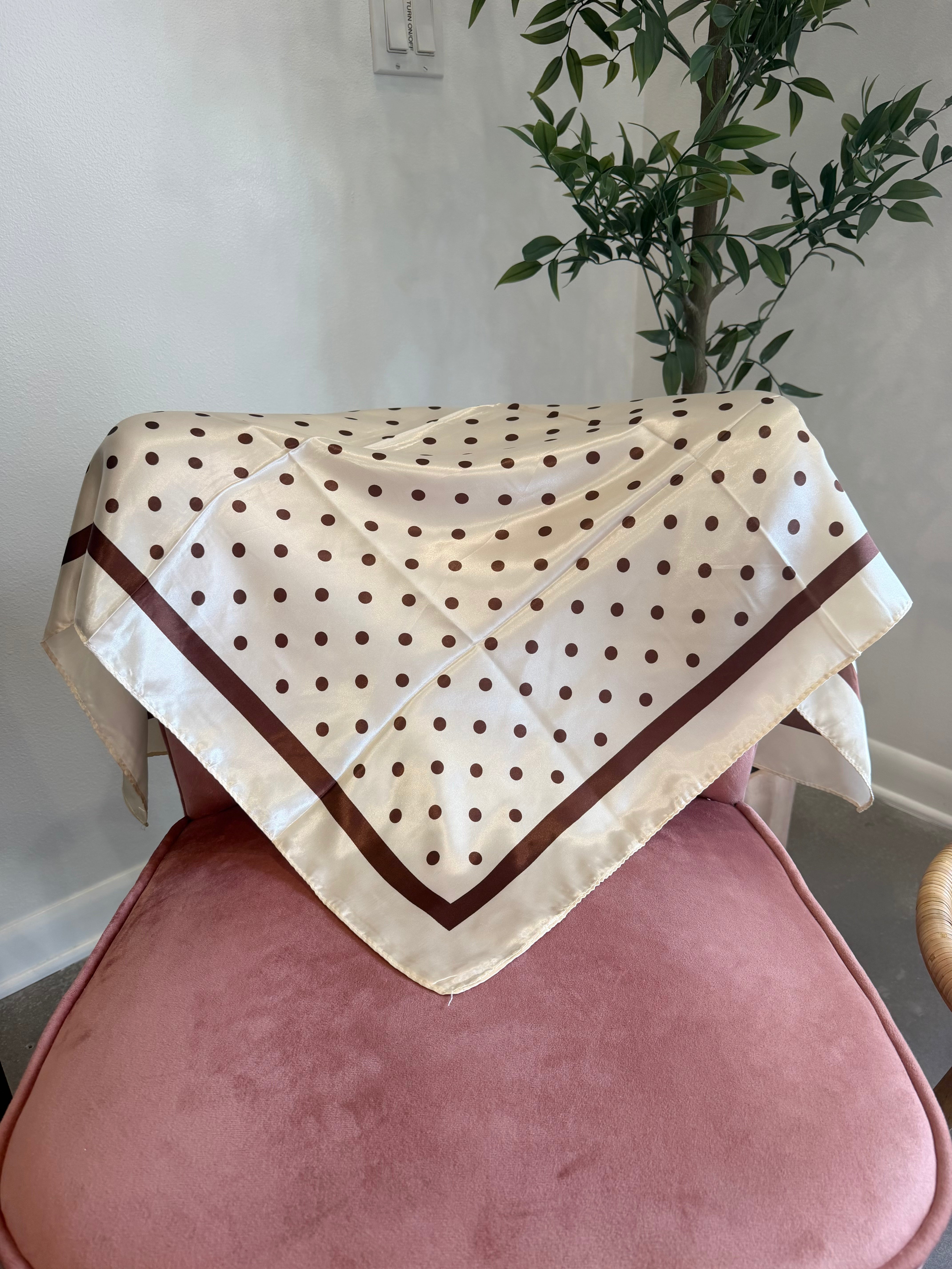 A cream-colored square scarf with brown polka dots and a brown border, draped over a dusty pink velvet chair in an indoor setting with a potted plant in the background.