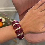 A person wearing a chunky, multi-band bracelet made of deep red resin with metallic gold accents, shown in a lifestyle setting with a pink chair in the background.