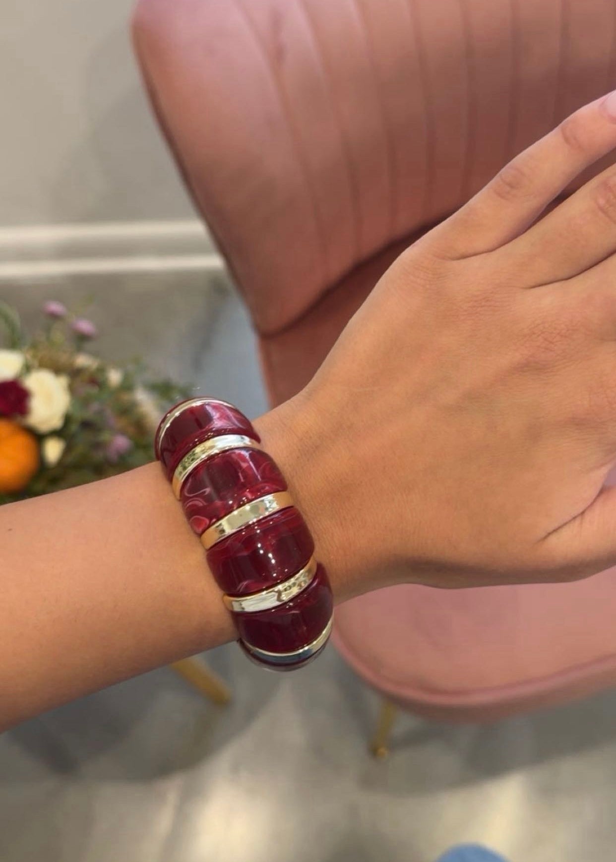 A person wearing a chunky, multi-band bracelet made of deep red resin with metallic gold accents, shown in a lifestyle setting with a pink chair in the background.