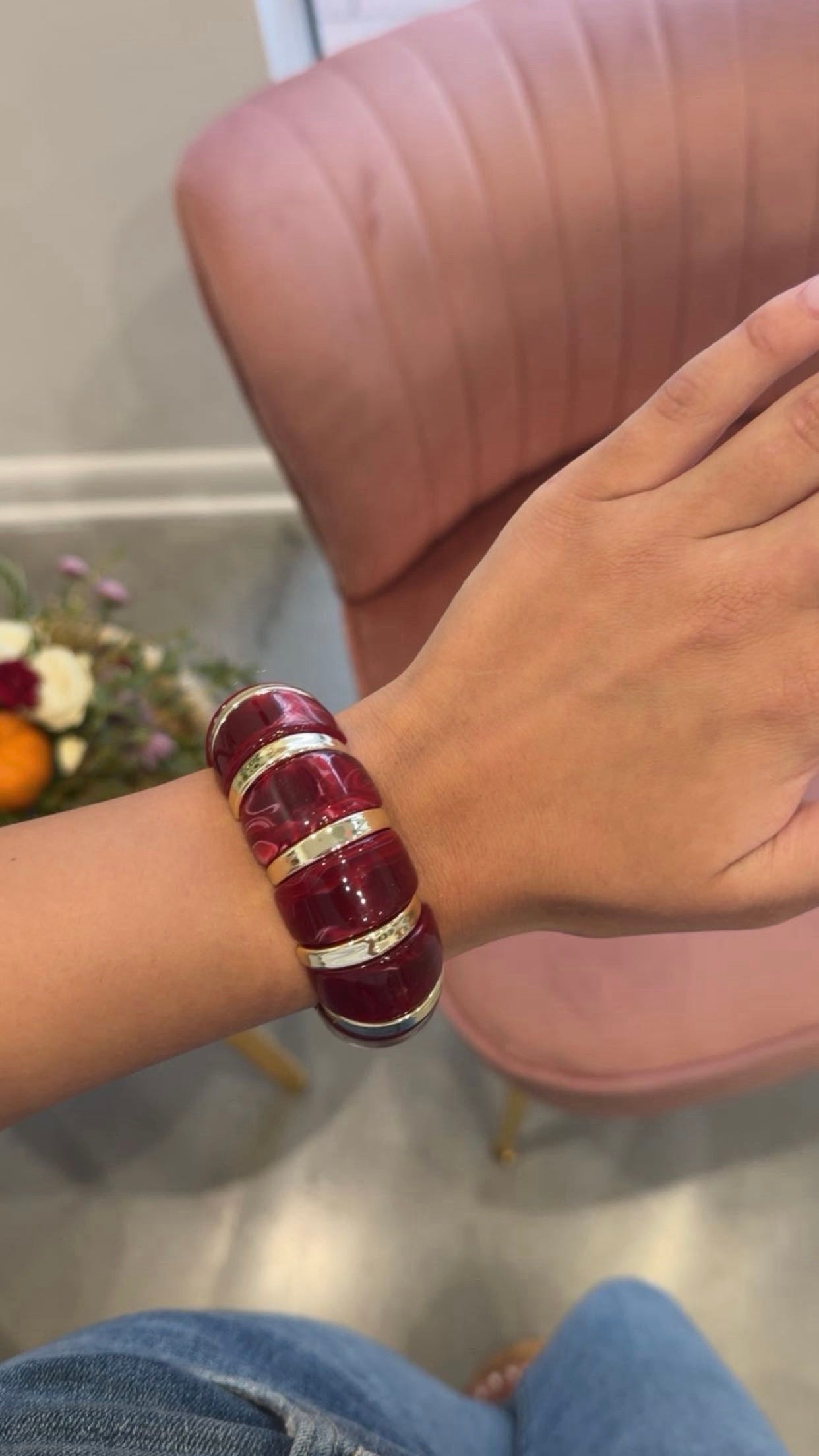 A person wearing a chunky, multi-band bracelet made of deep red resin with metallic gold accents, shown in a lifestyle setting with a pink chair in the background.