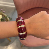A close-up of a person's wrist wearing a chunky, glossy deep red resin bangle bracelet with polished gold metal bands, set against a pink armchair in the background.