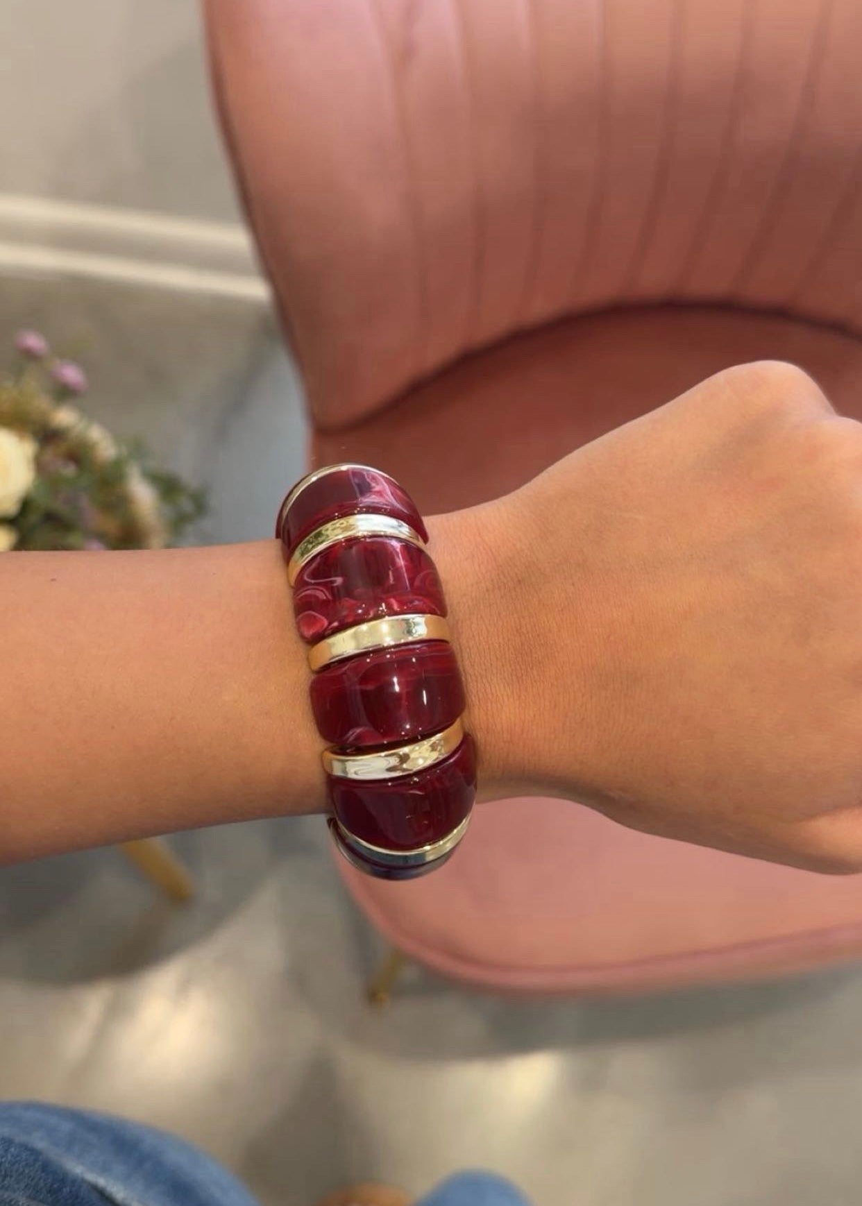 A close-up of a person's wrist wearing a chunky, glossy deep red resin bangle bracelet with polished gold metal bands, set against a pink armchair in the background.