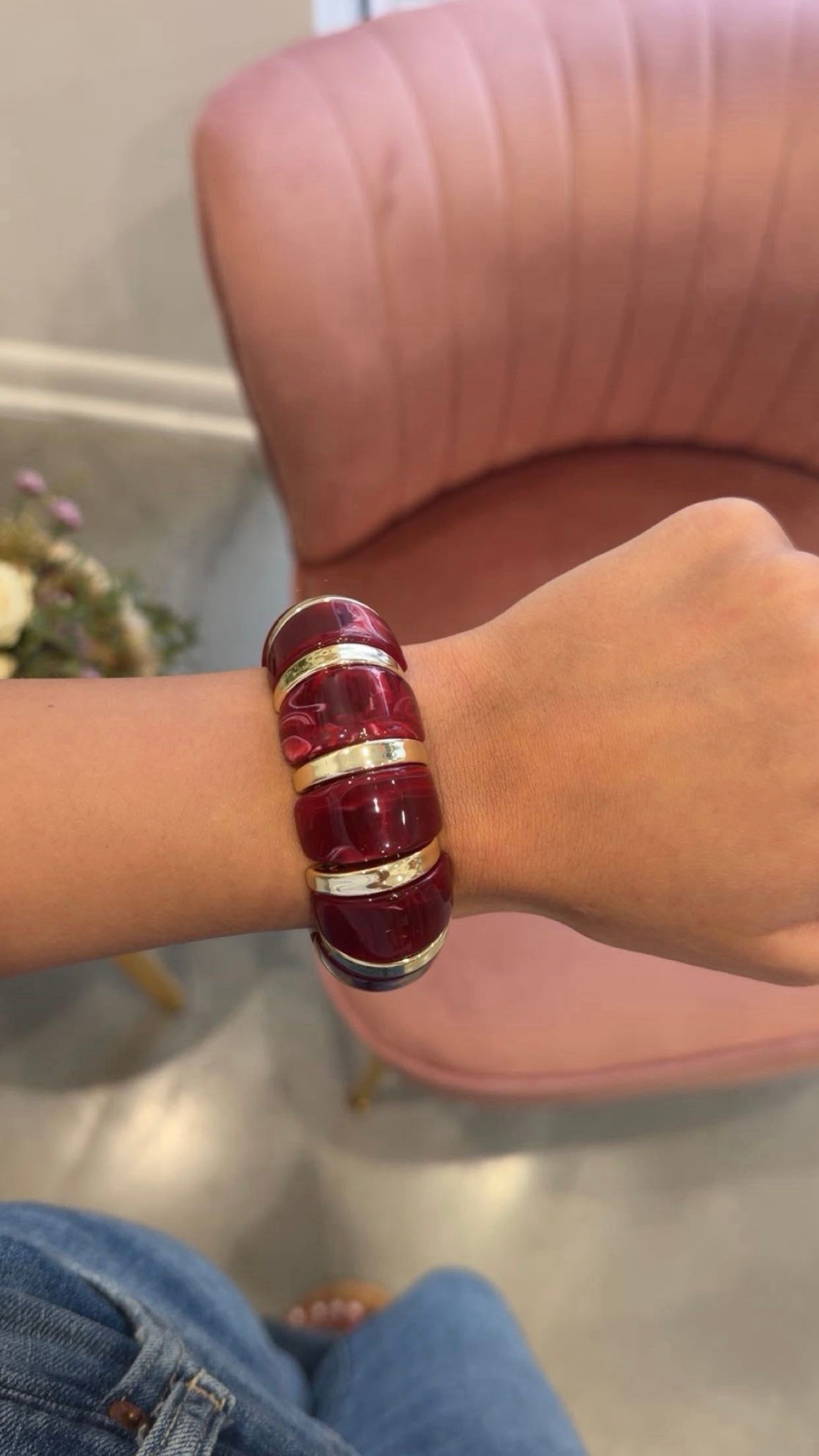 A close-up of a person's wrist wearing a chunky, glossy deep red resin bangle bracelet with polished gold metal bands, set against a pink armchair in the background.