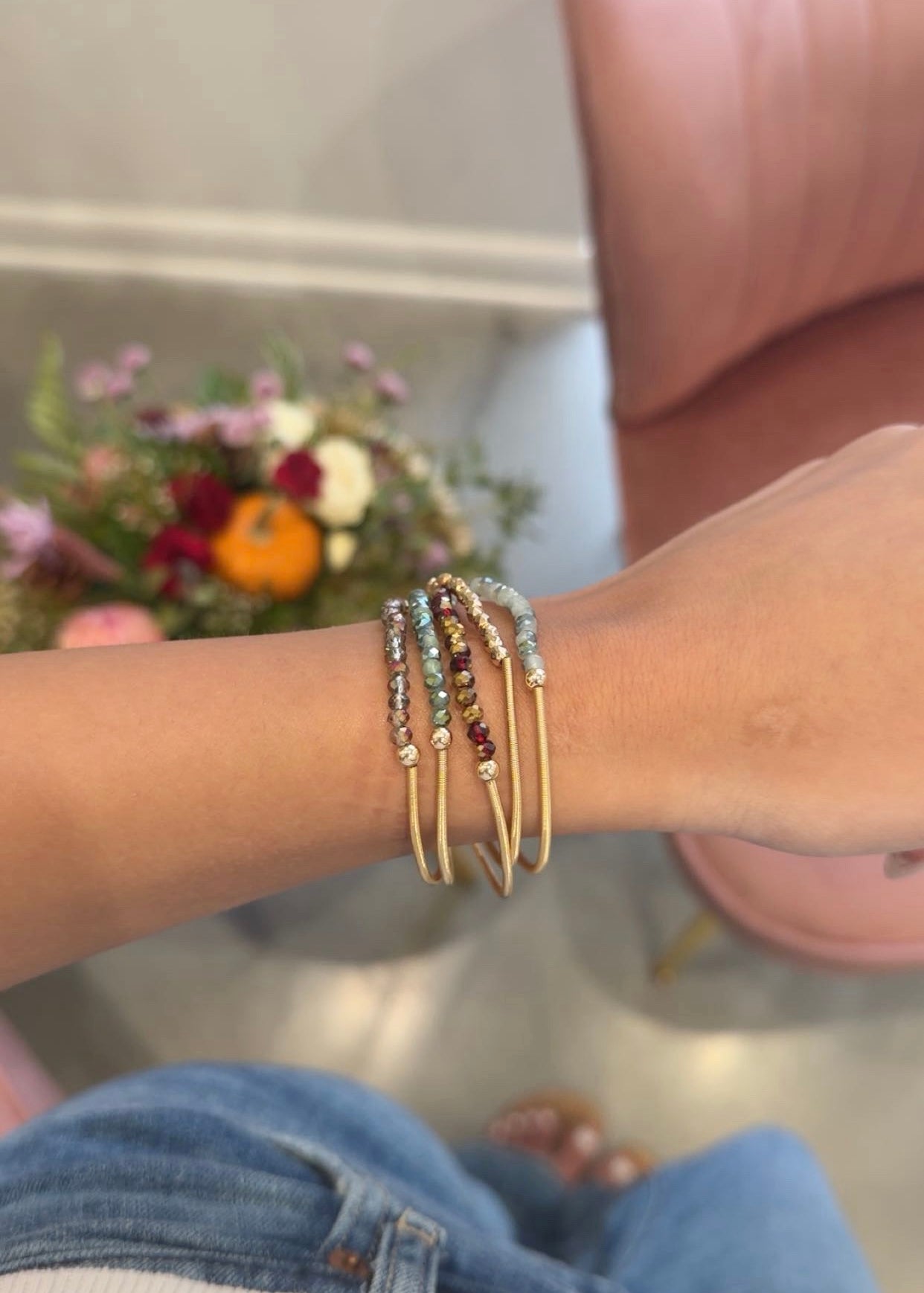A close-up of a person's wrist wearing a stack of gold bangle bracelets, some adorned with small multicolored beads. The person is wearing blue jeans, with a pink chair and a blurred floral arrangement in the background.