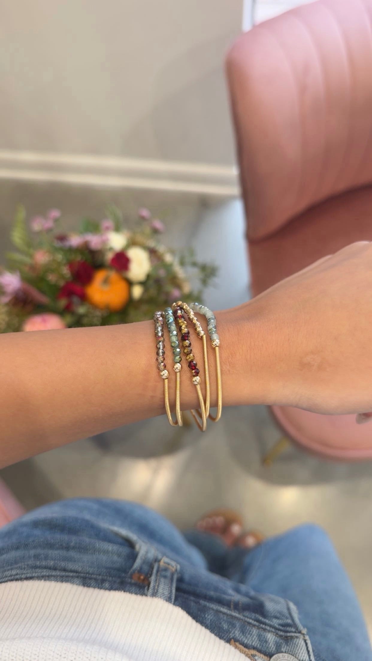 A close-up of a person's wrist wearing a stack of gold bangle bracelets, some adorned with small multicolored beads. The person is wearing blue jeans, with a pink chair and a blurred floral arrangement in the background.