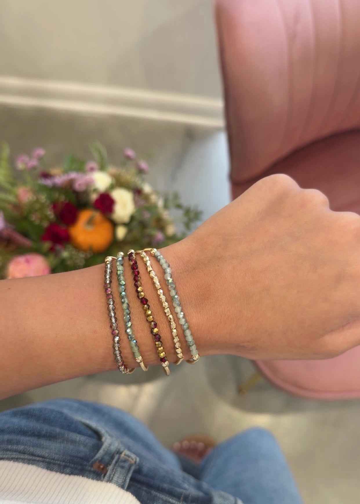 A close-up of a person's wrist wearing several delicate beaded bracelets, with a pink chair and flowers blurred in the background.