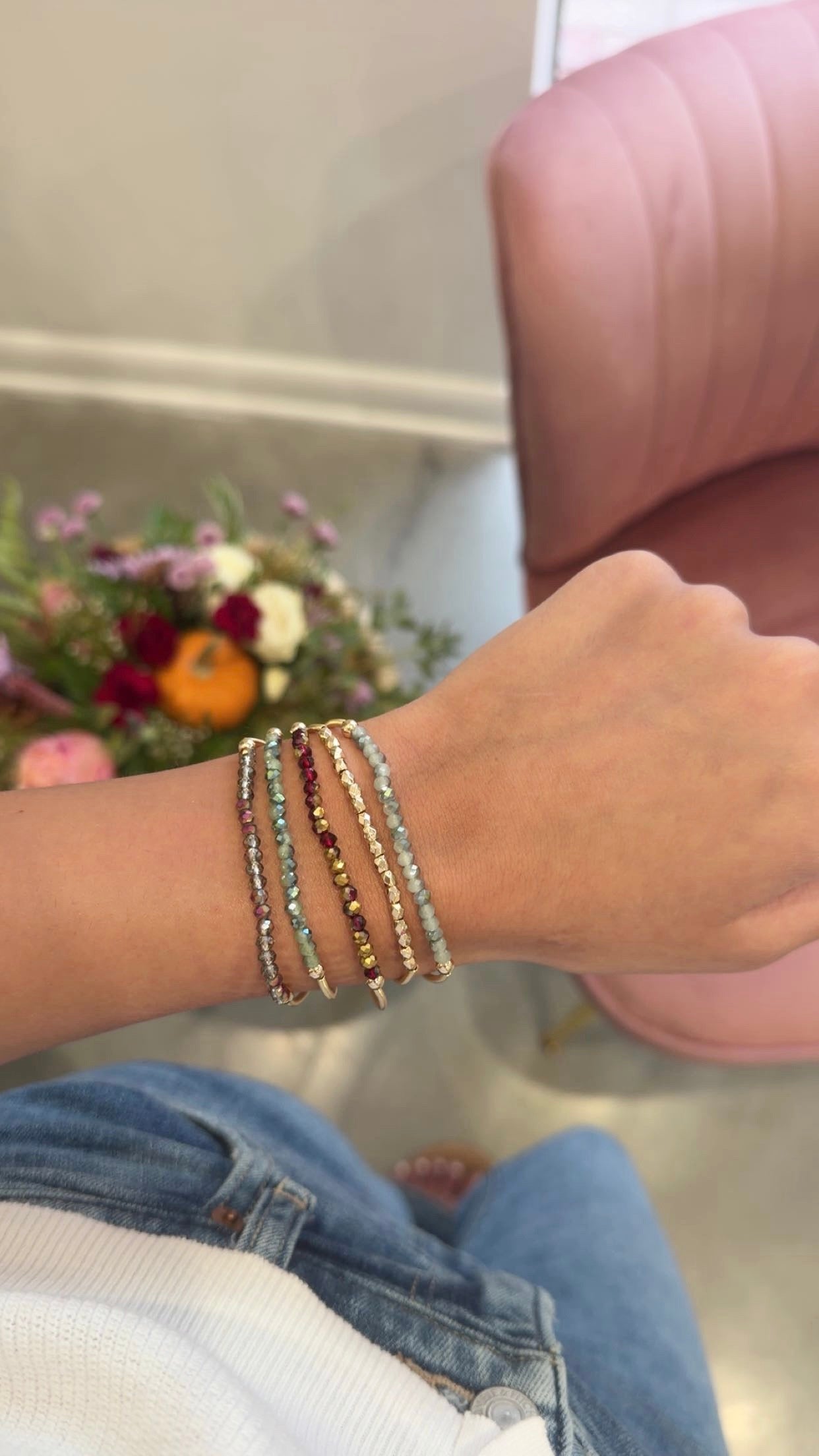 A close-up of a person's wrist wearing several delicate beaded bracelets, with a pink chair and flowers blurred in the background.