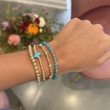 A close-up of a person's wrist wearing three beaded bracelets: two gold-toned with clear beads and one turquoise blue beaded bracelet with a small blue cross charm. The background shows a pink chair and a blurred floral arrangement.