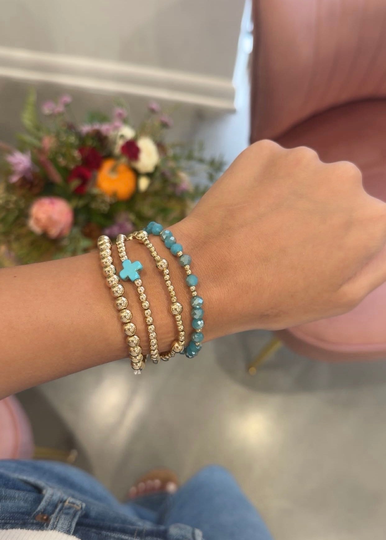 A close-up of a person's wrist wearing three beaded bracelets: two gold-toned with clear beads and one turquoise blue beaded bracelet with a small blue cross charm. The background shows a pink chair and a blurred floral arrangement.