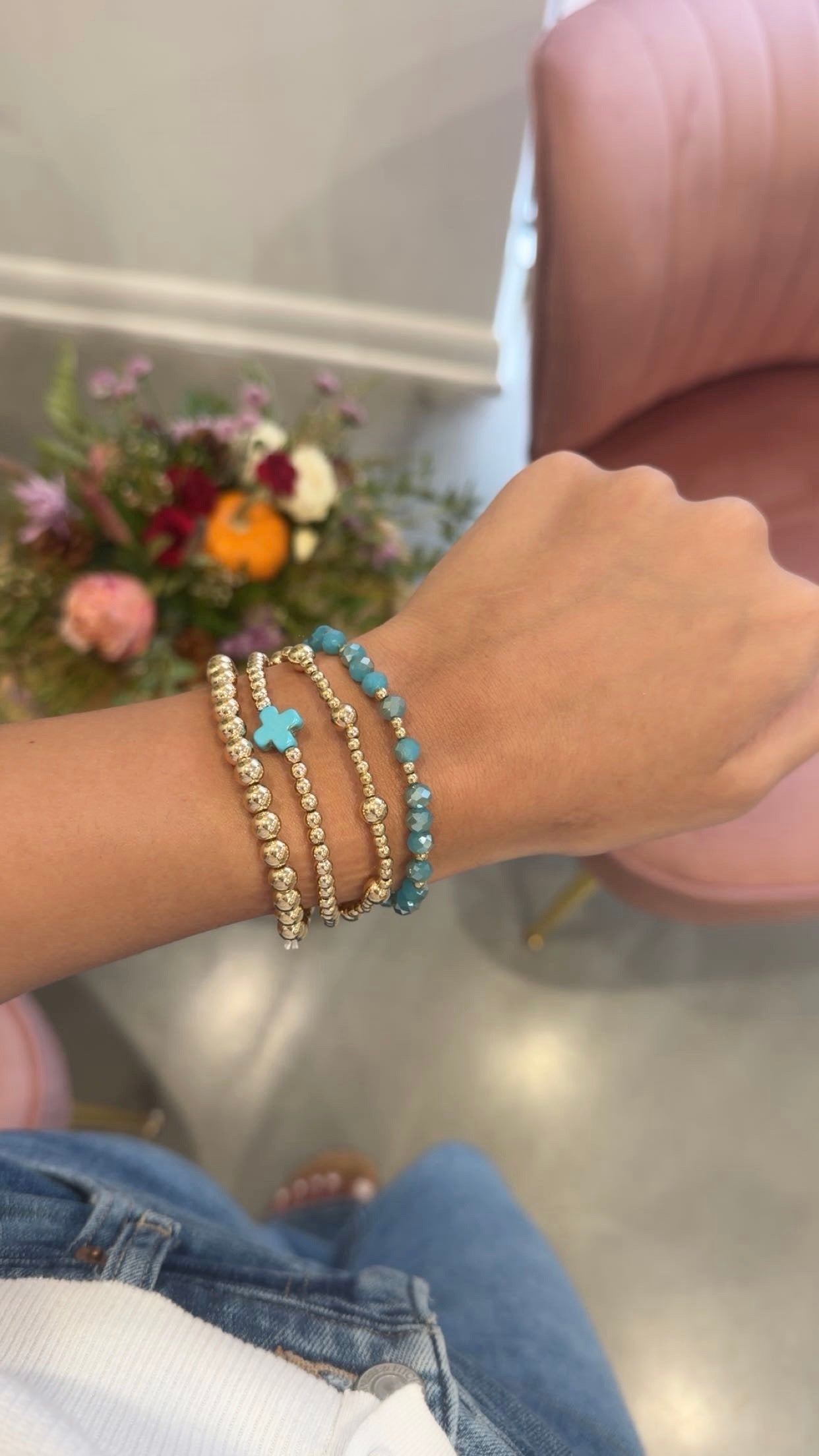 A close-up of a person's wrist wearing three beaded bracelets: two gold-toned with clear beads and one turquoise blue beaded bracelet with a small blue cross charm. The background shows a pink chair and a blurred floral arrangement.